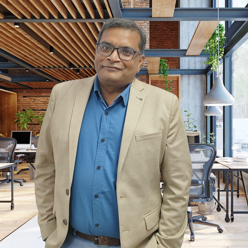 A confident man, presumably Ranjan Kumar, stands in a modern office environment, dressed in business casual attire with a warm smile.