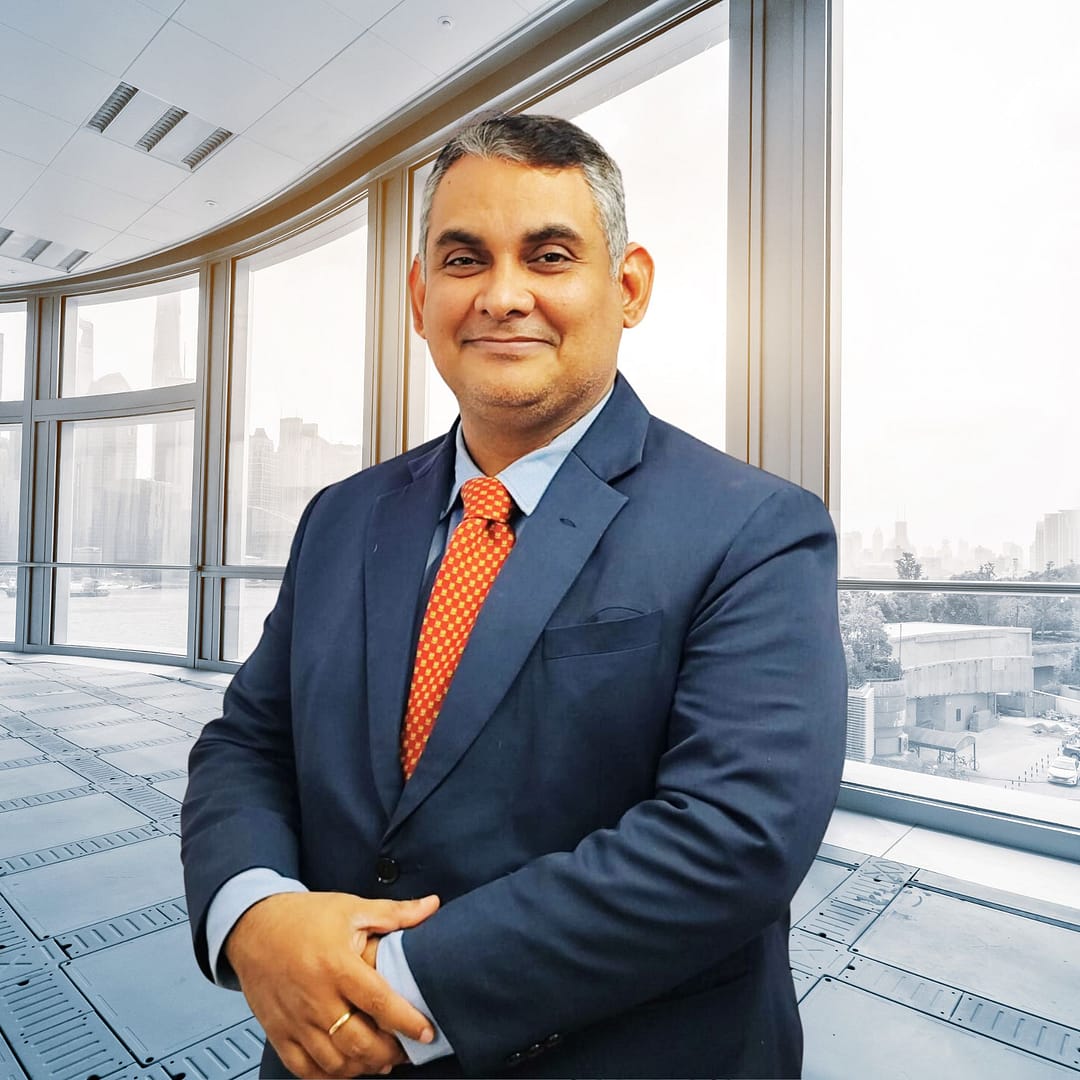 Praveen Kumar Mishra, dressed in a navy suit and orange tie, stands confidently in a high-rise office overlooking the city.
